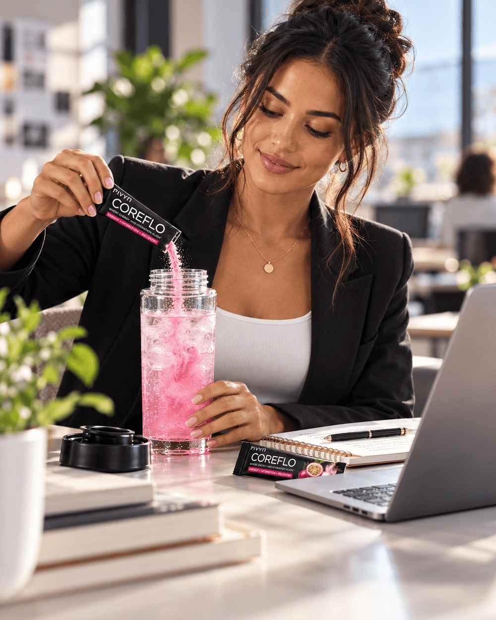 A woman in a black blazer and white top stands at a desk, pouring a pink drink from a clear plastic bottle into a black cup while holding a CoreFlo packet. A laptop, notebook, and plants are visible in a bright office setting.