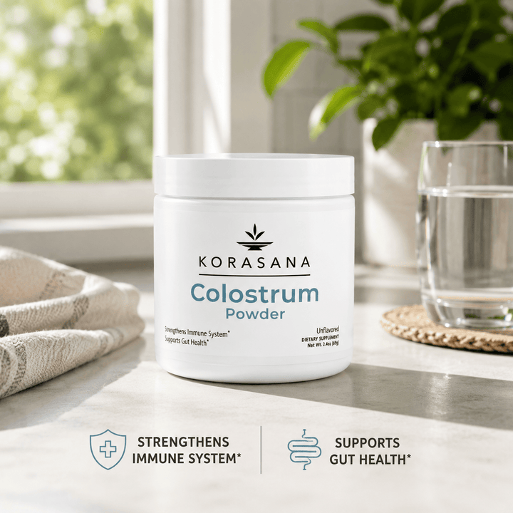 A white tub of KORASANA Colostrum Powder sits on a light countertop in a bright kitchen-like setting, with a glass of water, a folded towel, and a potted plant in the background. Blue text on the tub reads 'KORASANA Colostrum Powder,' and two feature badges at the bottom say 'Strengthens Immune System' and 'Supports Gut Health.
